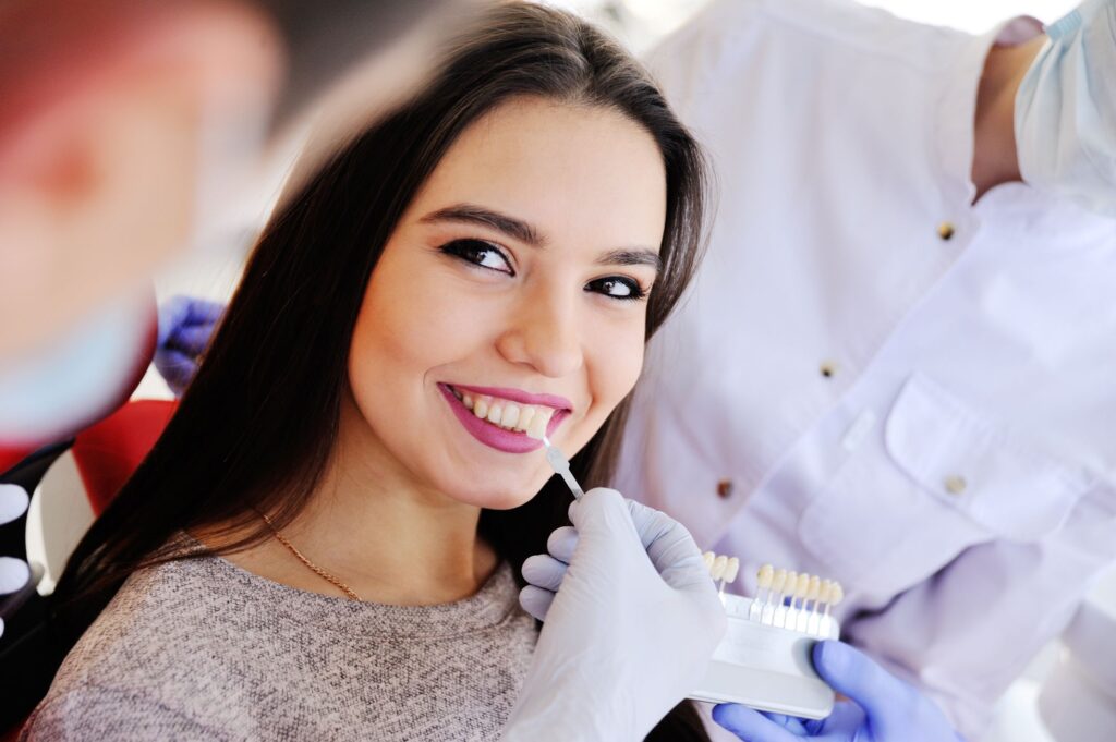 Woman with dark hair smiling at dentist holding shade guide to her teeth